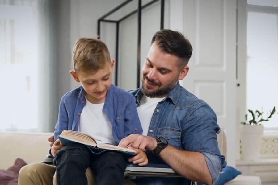 Disabled man sitting in wheelchair, with son on his lap reading a book.